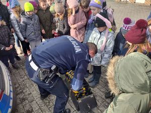 policjant prezentuje dzieciom wyposażenie radiowozu  w tle jezioro, zabudowania
