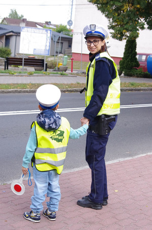 policjanci z przedszkolakami prowadzą działania na drodze w tle ulica i zabudowania