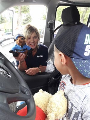 Chłopie siedzi w radiowozie na miejscu kierowcy,  w tle policjantka z maskotką