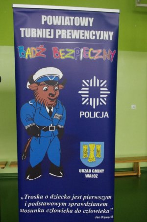 Maskotka policyjna, na niebieskim tle baneru, w tle nazwa turnieju i sentencja oraz telefon alarmowy