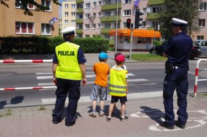 policjanci z 2 dzieci czekają na zmianę światła na sygnalizatorze, w tle budynki osiedla