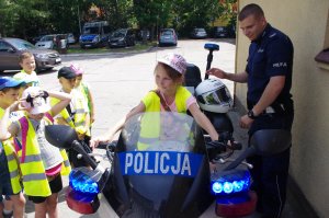 policjant stoi przy policyjnym motocyklu, na motocyklu siedzi dziewczynka w odblaskowej kamizelce, w tle inne dzieci