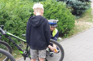 policjantka montuje odblask na rowerze, dziecko stoi tyłem,  w tle zarośla