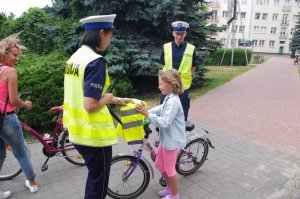 policjantka wręcza z policjantem kamizelkę odblaskową dziewczynce na rowerze, w tle zabudowania
