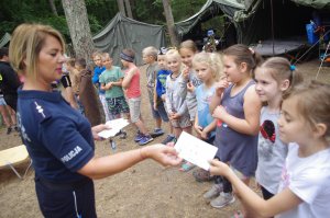policjantka wręcza kartki wakacyjne zuchom w tle obóz