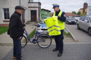 policjant drogówki wręcza mężczyźnie kamizelkę odblaskową,  w tle pojazdy i rower