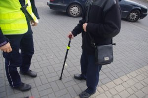 laska dla osoby niepełnosprawnej, którą trzyma mężczyzna, obok policjant z odblaskiem,  w tle pojazd