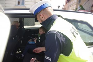 policjant wręcza odblask dziecku siedzącemu w aucie, w tle opiekun dziecka