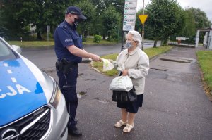 policjant wręcz seniorom opaski i torby, w tle radiowóz i parking samochodowy