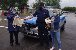 policjant wręcz seniorom opaski i torby, w tle radiowóz i parking samochodowy