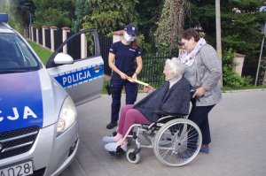 policjantka dzielnicowa wręcza gadżety seniorom w tle zabudowania i radiowóz