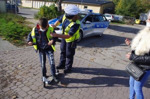 policjantka zakłada chłopcu z hulajnogą odblaskową kamizelkę, w tle zabudowania