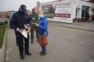 policjant wręcza seniorom kobietom i mężczyznom torby na zakupy, opaski odblaskowe w tle zabudowania i zarośla