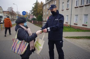 policjant wręcza seniorom kobietom i mężczyznom torby na zakupy, opaski odblaskowe w tle zabudowania i zarośla
