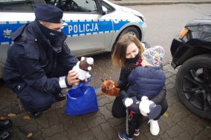 policjanci wręczają dzieciom maskotki, obok rodzice w tle zabudowania i radiowóz