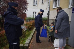 policjanci wręczają maskotki, przy dzieciach rodzice i opiekunowie w tle zabudowania