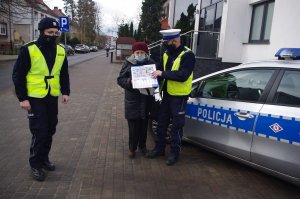 policjanci stoją przy radiowozie, w środku kobieta trzyma otrzymany kalendarz, w tle chodnik i ulica