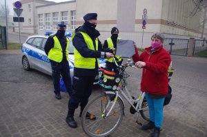 policjant wręcza rowerzystce torbę odblaskową, w tle policjanci i zabudowania