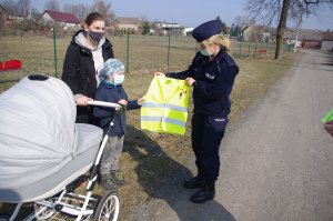 policjanci wręczają odblaski kobiecie z wózkiem i dzieckiem,w tle zabudowania