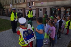 policjantka przedstawia dzieciom kierowanie ruchem,  w tle budynek szkoły