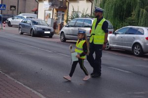 dziewczynka pod opieką policjanta przechodzi przez drogę,  w tle pojazdy