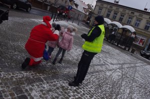 Mikołaj częstuje dzieci cukierkami obok policjantka w tle miasto