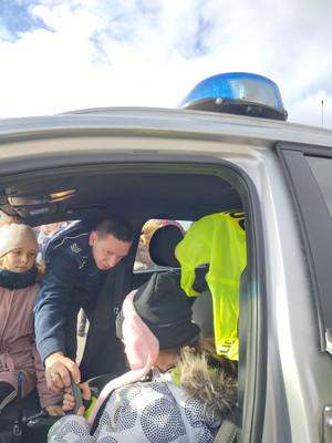 policjant prezentuje dzieciom wyposażenie radiowozu  w tle jezioro, zabudowania