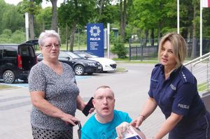 policjantka, osoba niepełnosprawna na wózku inwalidzkim, matka w tle pojazdy, parking i logo Policji