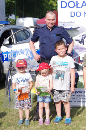 policjant rozmawia z dziećmi w tle policyjny baner i radiowóz