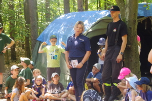 służby rozmawiają z uczestnikami w tle las i namioty
