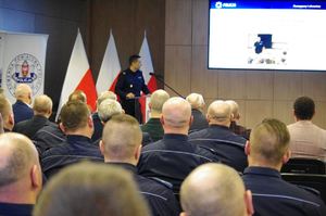 policjanci i zaproszeni goście siedzą w sali w tle ściany sali i banery jednostki oraz trzy stojące flagi Polski
