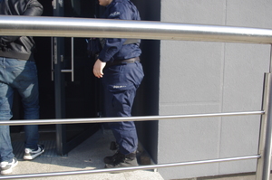 policjant z zatrzymanym  w tle drzwi wejściowe do budynku komendy