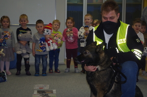 policjanci wraz z psem policyjnym rozmawiają z dziećmi w tle sala w przedszkolu
