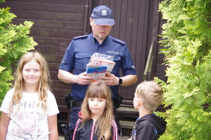 policjant wręcza dzieciom niespodzianki w tle dom drewniany Brda
