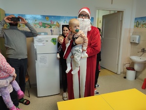 Mikołaj w sali rozdaje prezenty w tle policjanci, strażacy i dzieci