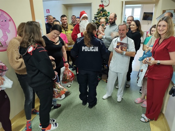 Mikołaj w sali rozdaje prezenty w tle policjanci, strażacy i dzieci