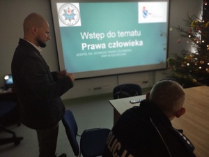 policjant w cywilnym ubraniu prowadzi spotkanie w tle sala i ekran a na nim film