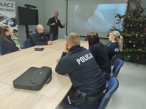 policjant w cywilnym ubraniu prowadzi spotkanie w tle sala i ekran a na nim film