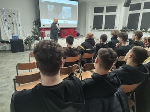 policjant w cywilnym ubraniu prowadzi spotkanie w tle sala i ekran a na nim film