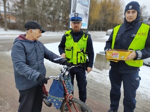 policjanci wręczają seniorom odblaskowe opaski i częstują ciastkami
