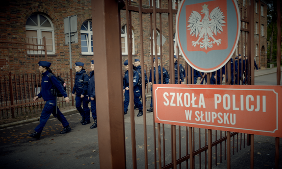 brama wejściowa do Szkoły Policji w Słupsku w tle idący policjanci i mury budynku szkoły