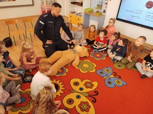 policjant z użycie misia uczy dzieci udzielania pomocy poszkodowanym w tle sala i siedzące na dywanie dzieci