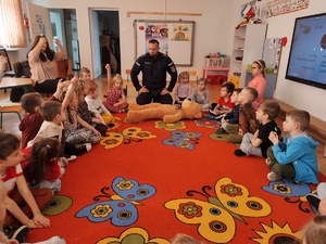 policjant z użycie misia uczy dzieci udzielania pomocy poszkodowanym w tle sala i siedzące na dywanie dzieci