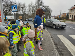 policjanci zabezpieczają bezpieczeństwo na drodze, a dzieci w towarzystwie pluszowego żubra przechodzą po przejściu w tle ulica i pojazdy