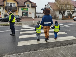 policjanci zabezpieczają bezpieczeństwo na drodze, a dzieci w towarzystwie pluszowego żubra przechodzą po przejściu w tle ulica i pojazdy