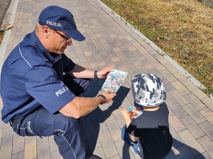 policjant wręcza dziecku kolorowankę w tle droga i zabudowania