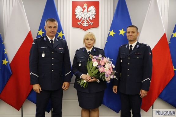 policjantka w otoczeniu dwóch policjantów w tle godło, w tle flaga Polski i Unii Europejskiej