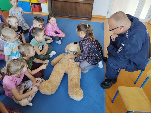 policjanci uczą dzieci zasad udzielania pomocy. W tle sala przedszkolna i dzieci