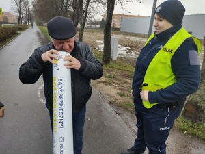 policjantka i strażnik gminny prezentują odblaski pieszym i rowerzystom w tle ruch uliczny i zabudowania