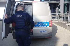 policjant otwiera tylne drzwi radiowozu w tle budynek komendy